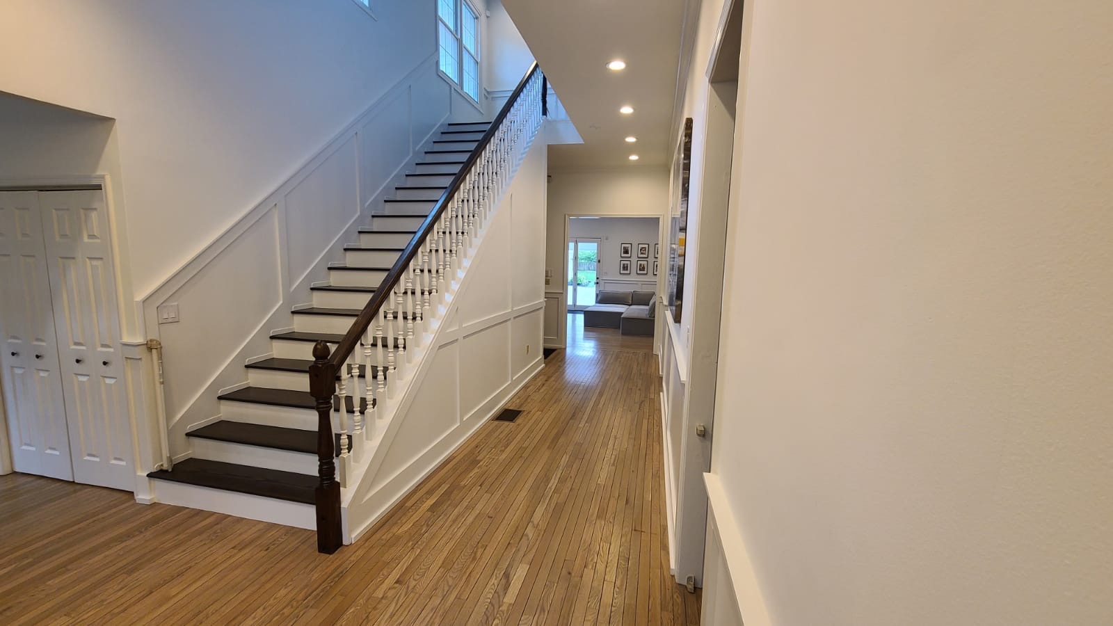 Bright hallway with hardwood floors, white wainscoting, and a staircase with dark wood steps and railing leading to a second floor, opening into a living area.