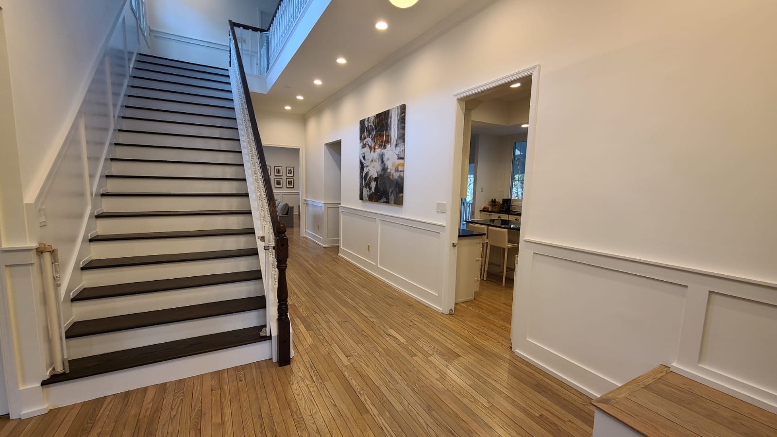Spacious hallway with hardwood floors, white wainscoting, dark wood staircase, recessed lighting, abstract wall art, and view into a kitchen and living area.