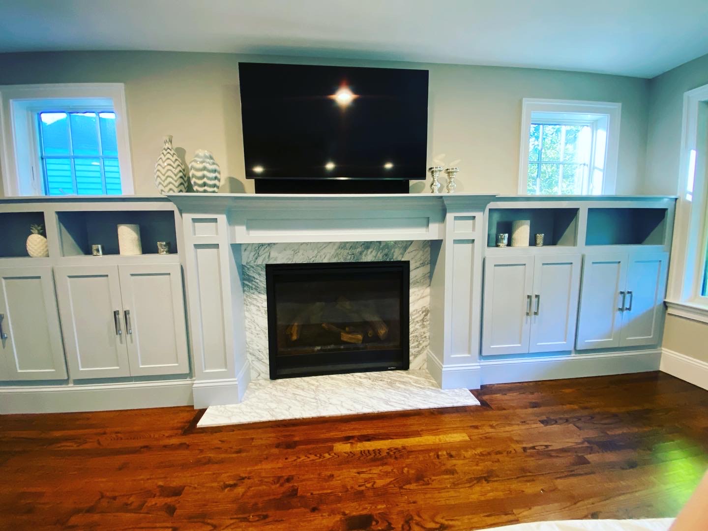 Living room with built-in gray cabinetry, marble fireplace, mounted TV, decorative vases, candles, and hardwood floors beneath large windows.