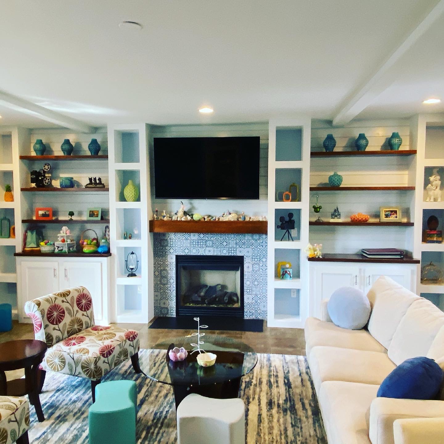 Colorful living room with built-in shelving, TV above a tiled fireplace, dark wood mantel, patterned armchairs, glass coffee table, and neutral sofa with pillows.