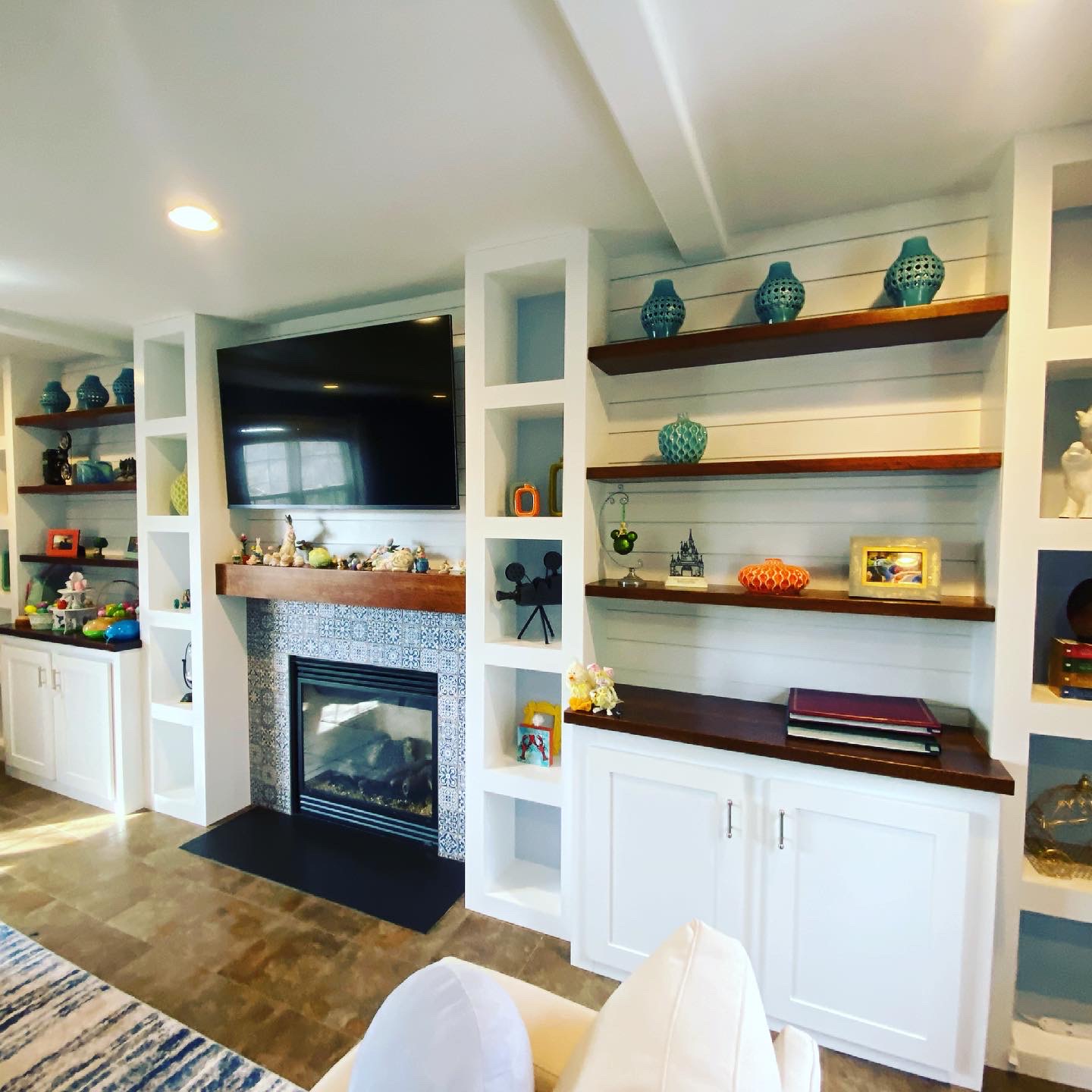 Living room with built-in shelving, dark wood accents, shiplap backing, colorful decor, TV above a tiled fireplace, and white cabinetry with silver handles.