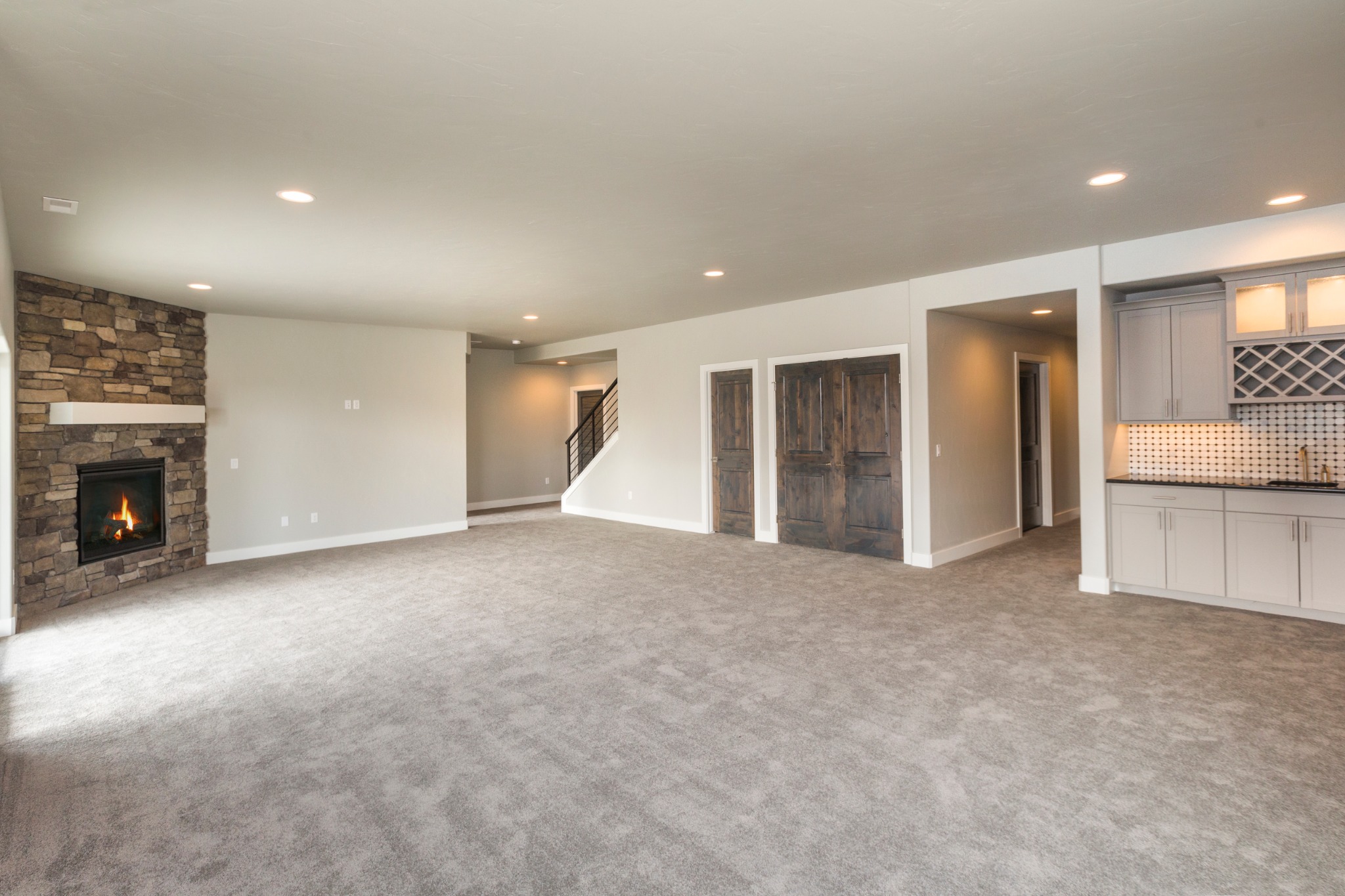 Spacious basement with gray carpet, stone fireplace, built-in wet bar with wine rack, wood doors, and recessed lighting creating a cozy, modern ambiance.