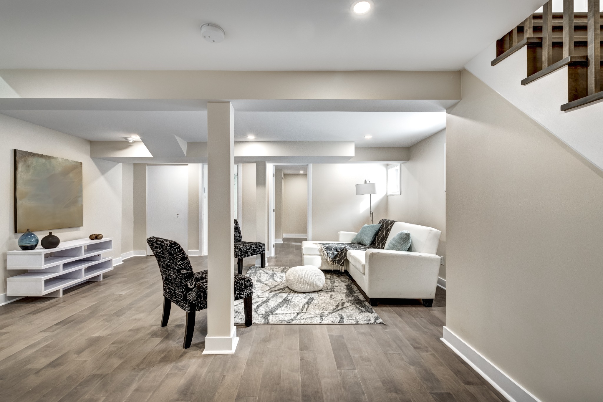 Modern finished basement with light wood flooring, neutral walls, cozy seating area with a white sofa, accent chairs, and decorative art and lighting.