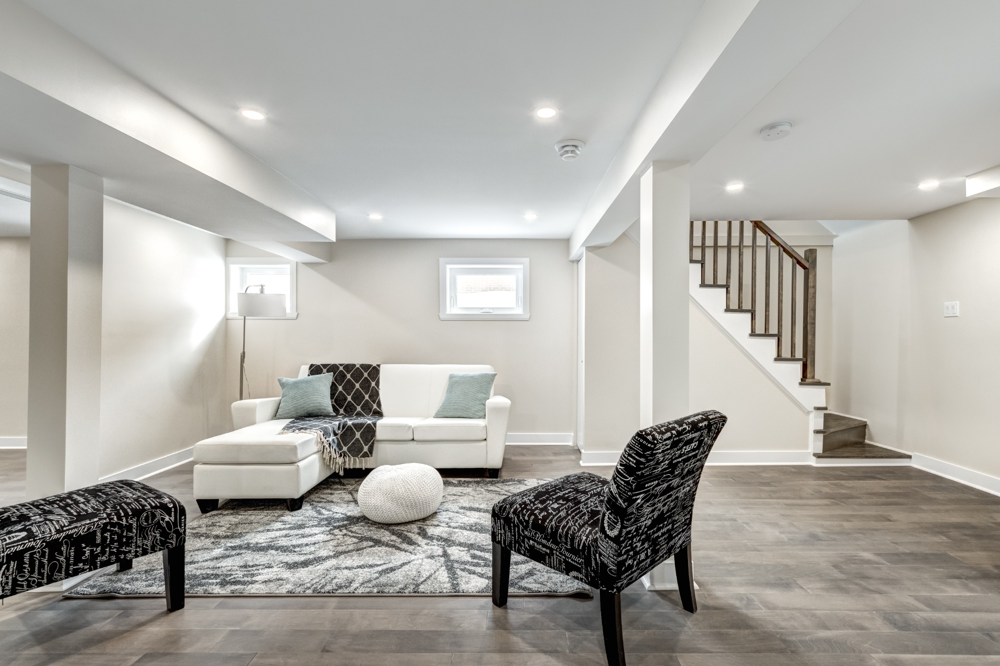 Bright basement living area with light wood floors, white sectional sofa, patterned black chairs, soft rug, and stairs with a wooden handrail in the corner.
