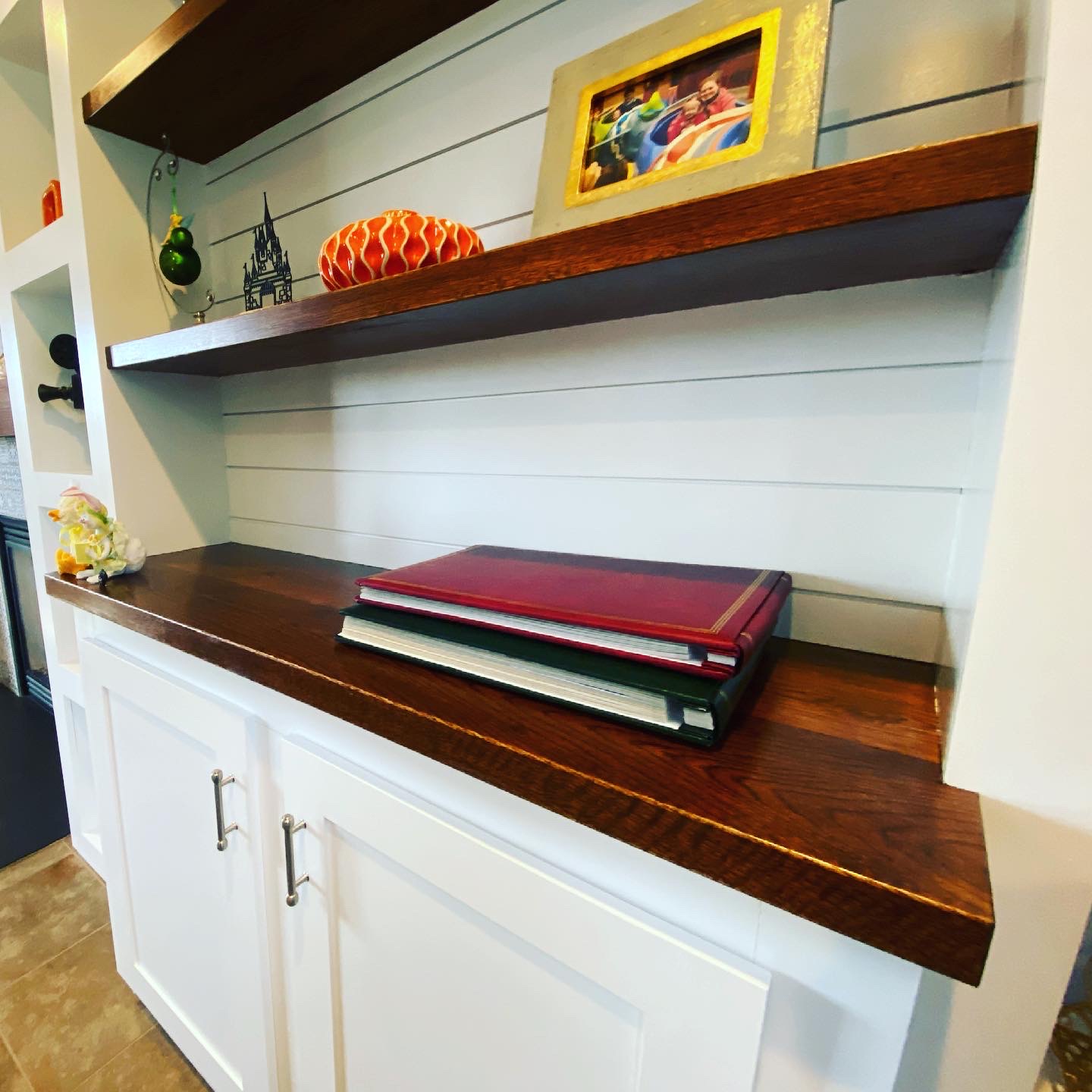 Built-in shelving with dark wood shelves, white shiplap backing, photo albums, framed photo, decorative items, and white cabinetry with silver handles.