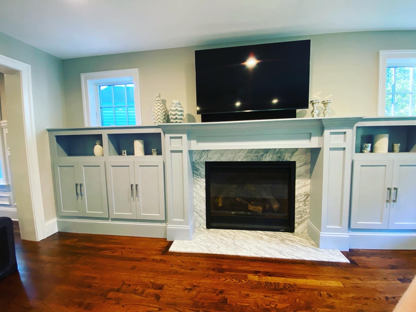 Built-in gray cabinetry with a marble-surround fireplace, mounted TV above, decorative vases, candles, and hardwood flooring in a cozy living space.