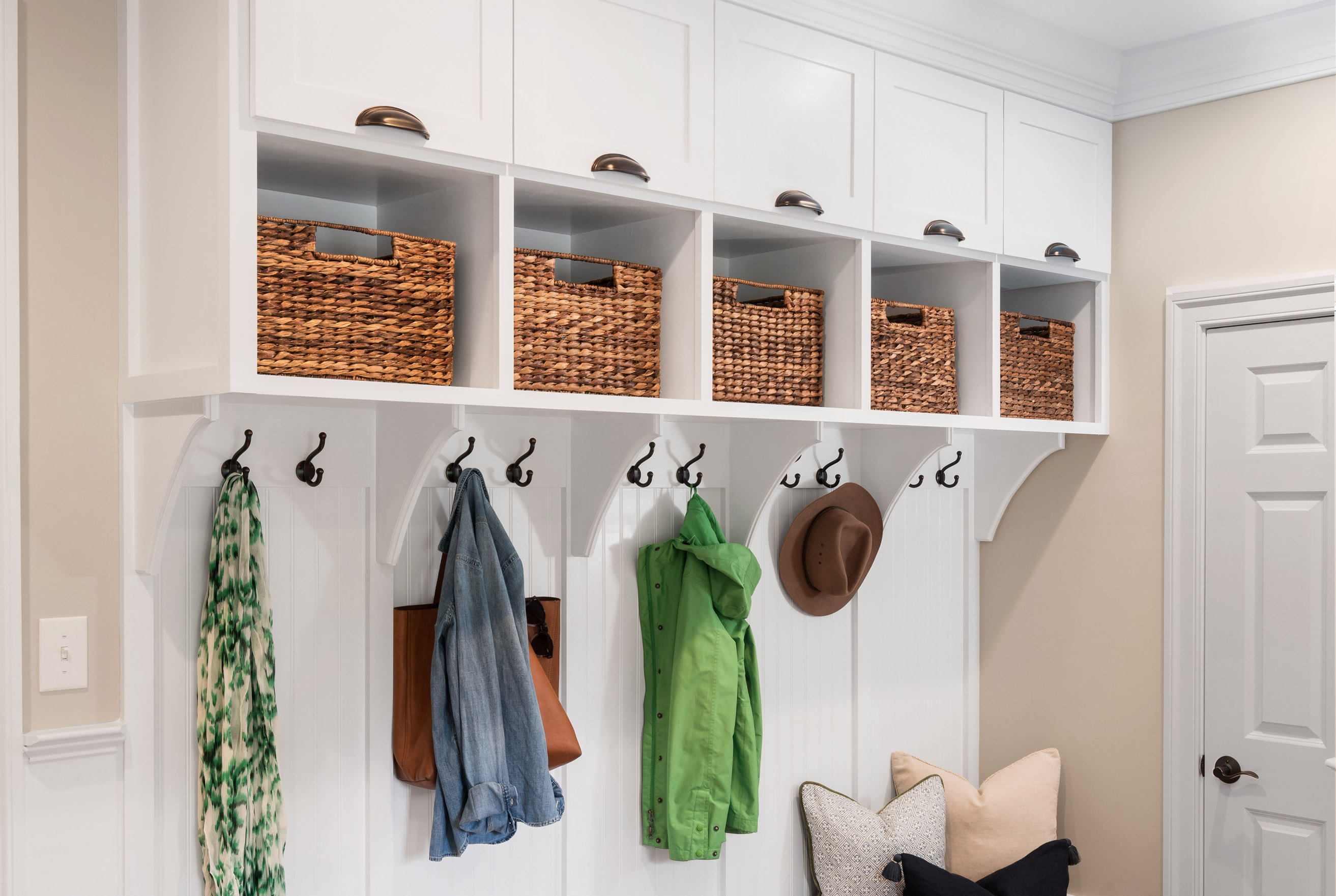 White mudroom storage with upper cabinets, cubbies holding woven baskets, coat hooks with jackets and hats, and a bench with pillows below.