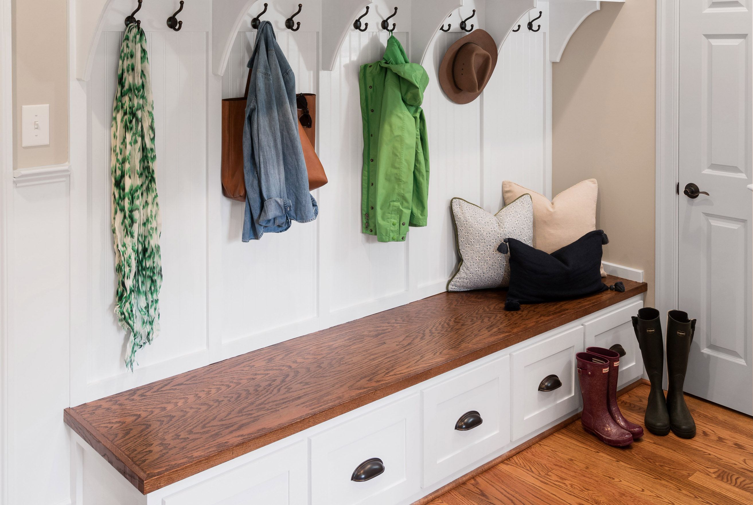 Mudroom with white built-in storage, dark wood bench, coat hooks holding jackets and hats, decorative pillows, and rain boots on hardwood floor.