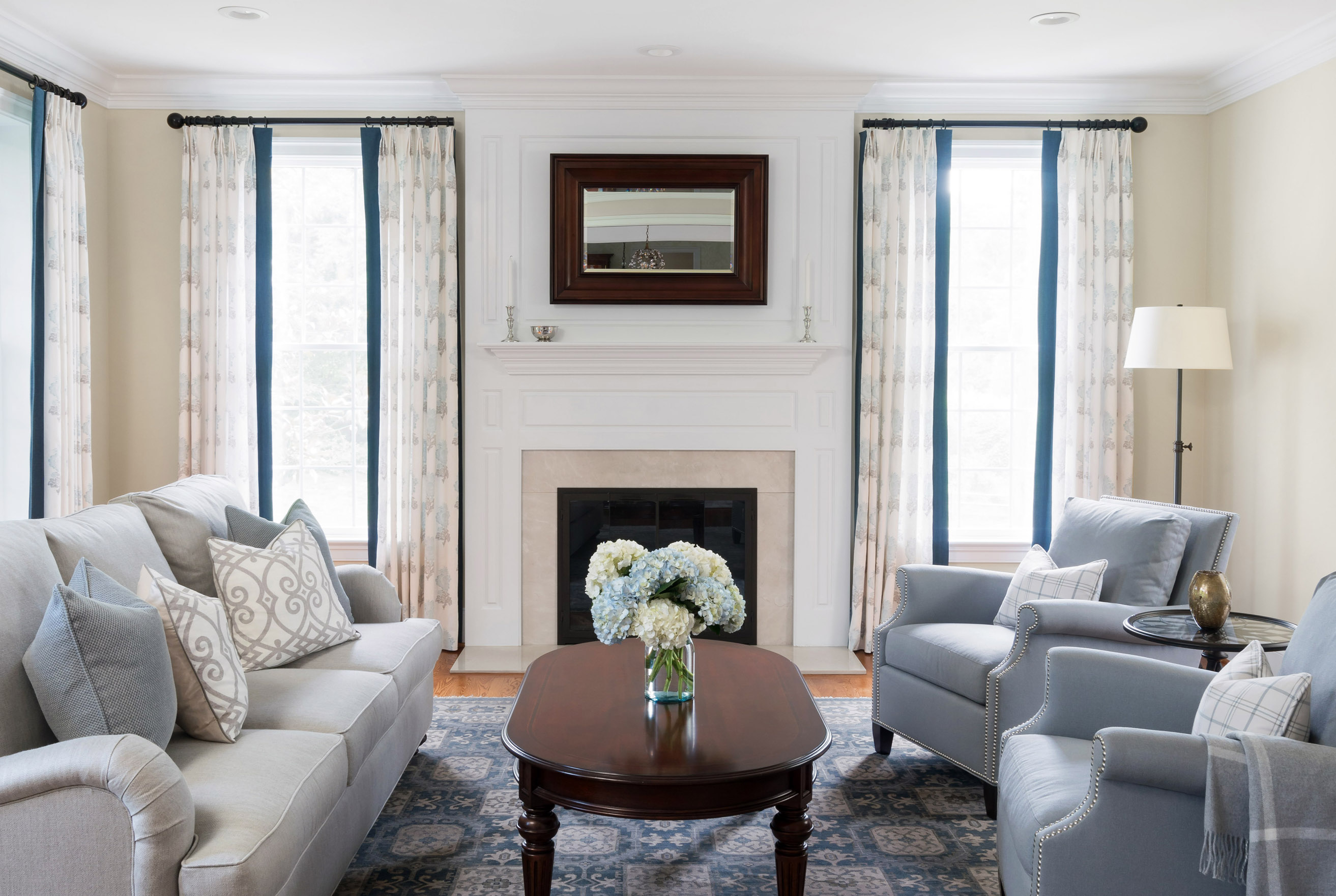 Elegant living room with light gray sofa and chairs, dark wood coffee table, floral centerpiece, fireplace with mirror above, and tall windows with curtains.