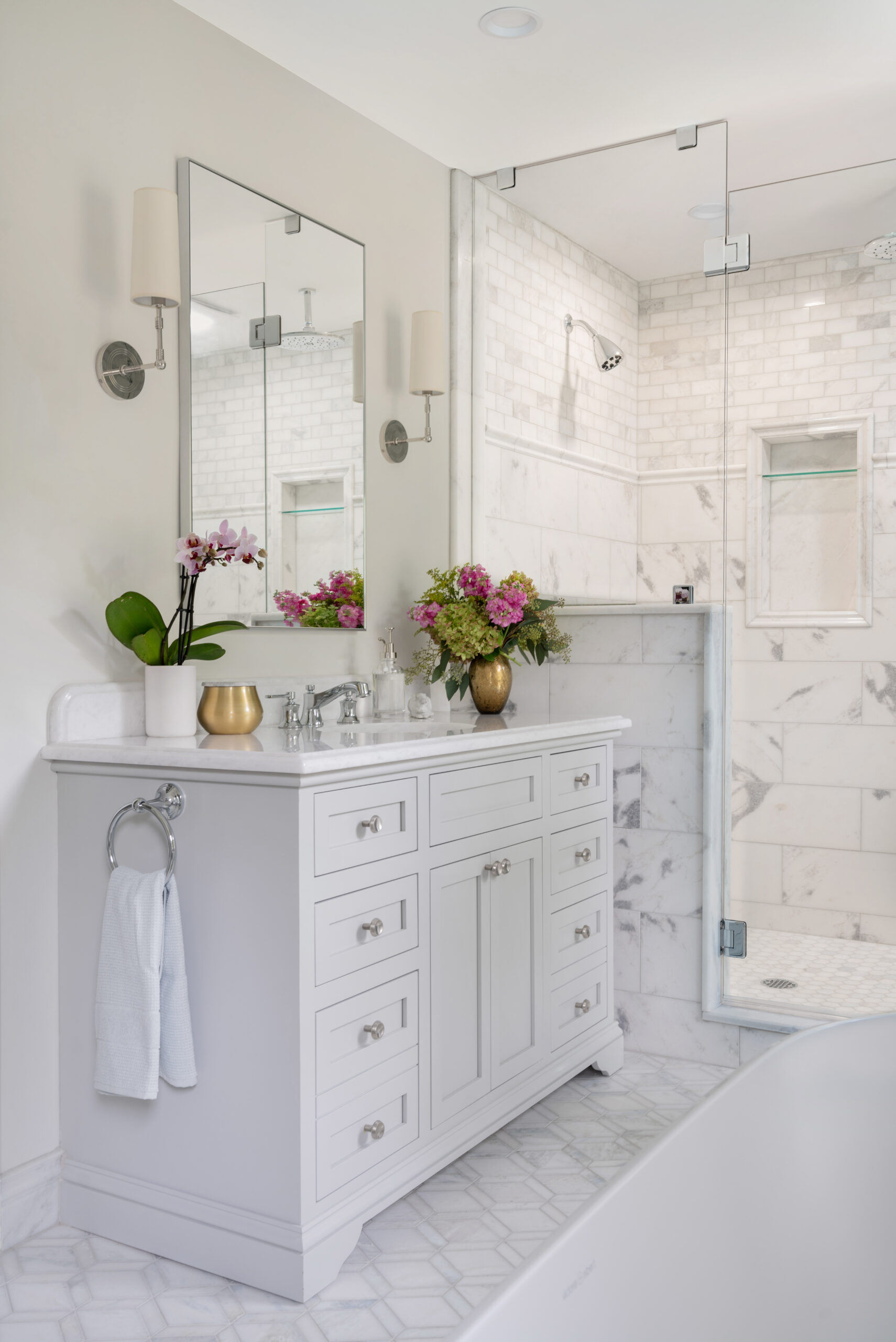 Elegant bathroom with white vanity, marble countertop, floral decor, large mirror, wall sconces, glass-enclosed marble shower, and geometric tile floor.