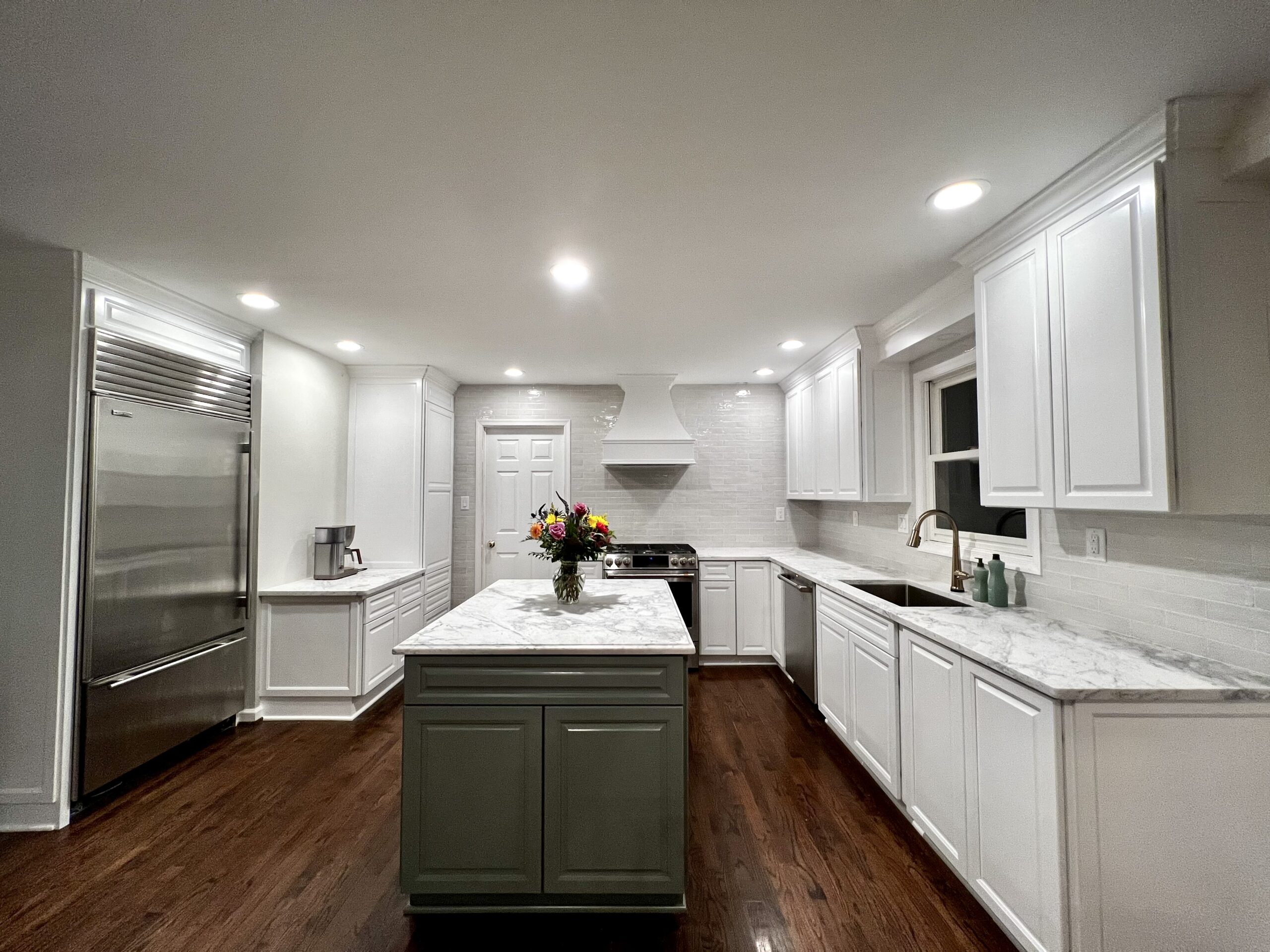 Bright kitchen featuring white cabinetry, marble countertops, a gray island with flowers, stainless steel appliances, and dark hardwood floors.