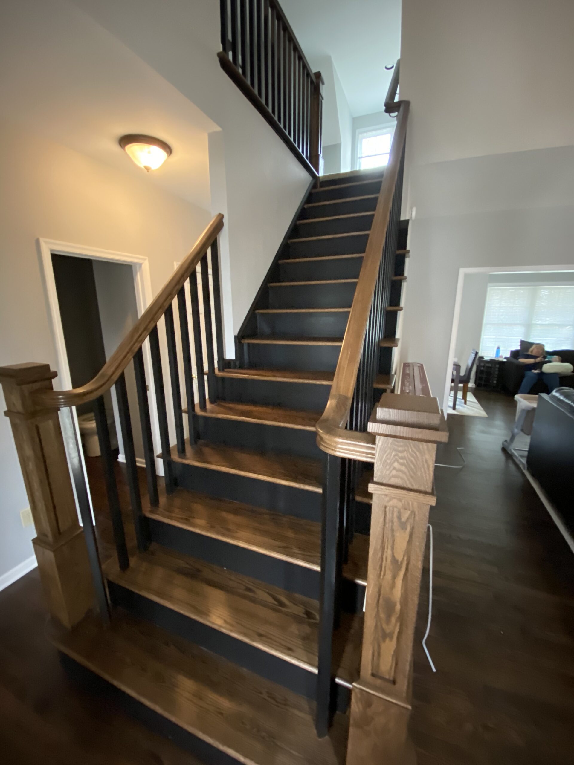 Wooden staircase with dark treads, black risers, and black spindles, featuring a warm wood handrail and newel posts in a modern, open hallway.