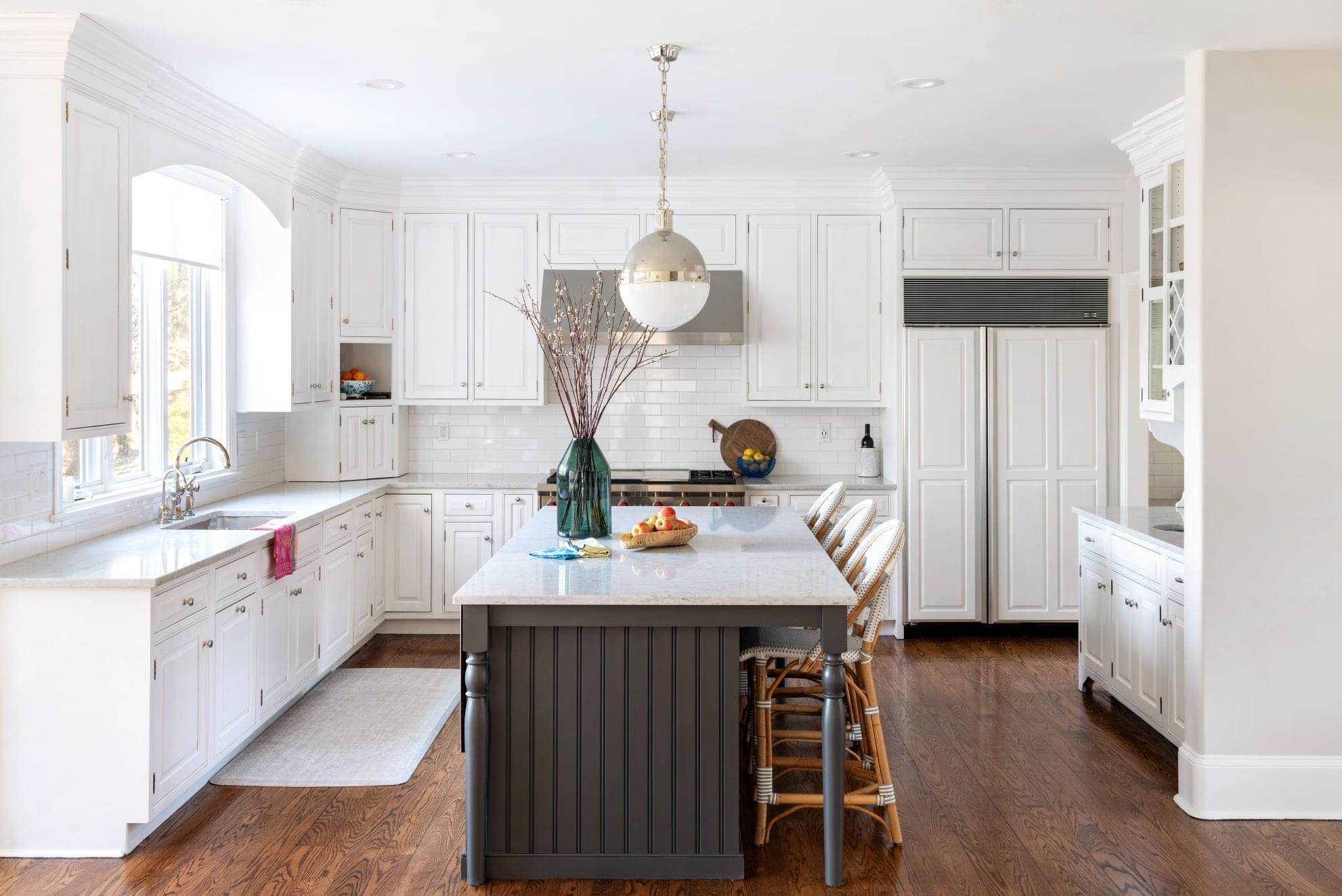 Bright kitchen with white cabinetry, subway tile backsplash, dark gray island with marble countertop, rattan barstools, wood flooring, and modern pendant light.