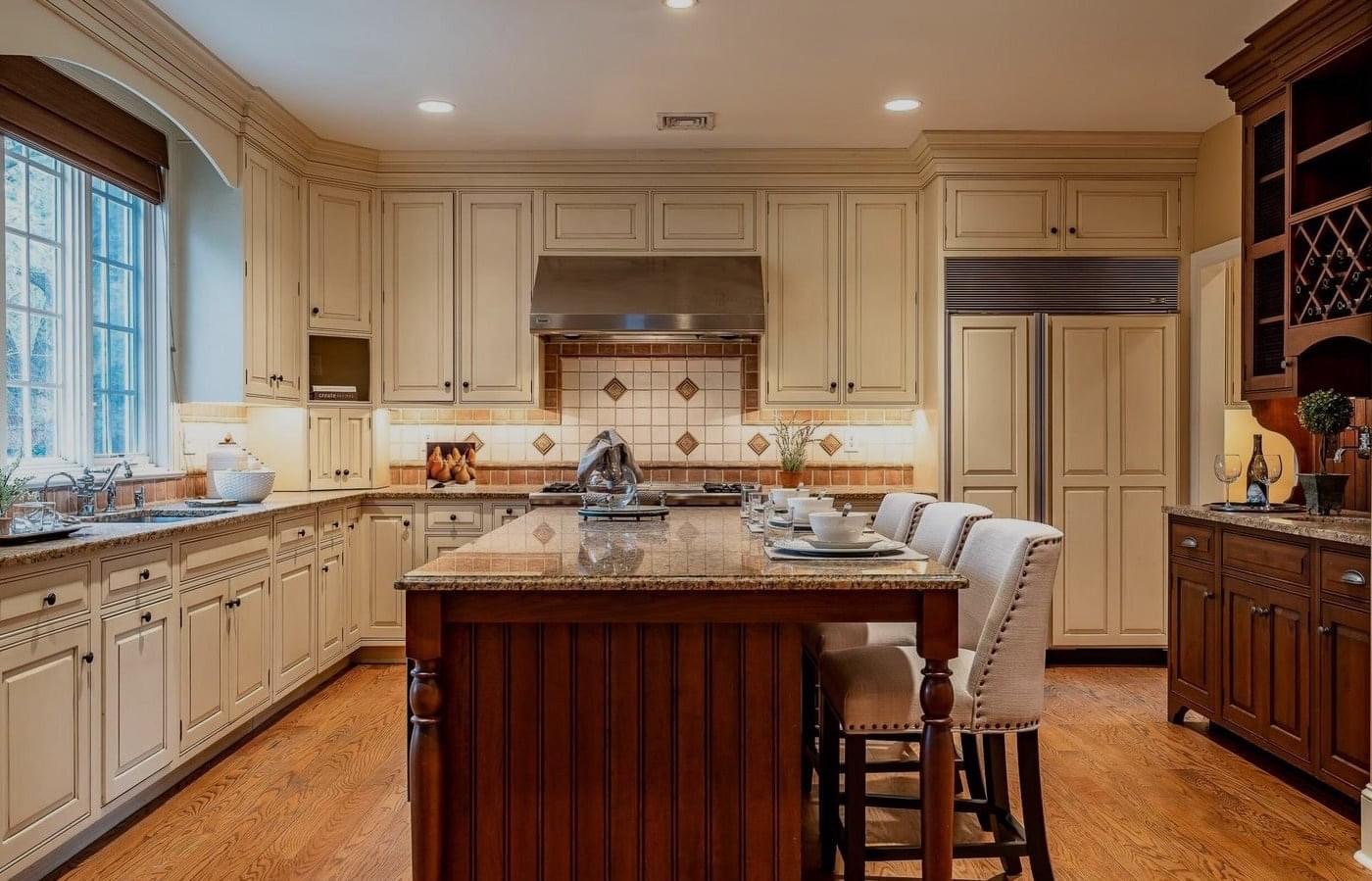 Warm traditional kitchen with cream cabinetry, granite countertops, tile backsplash, wood island with seating, stainless steel range hood, and hardwood floors.