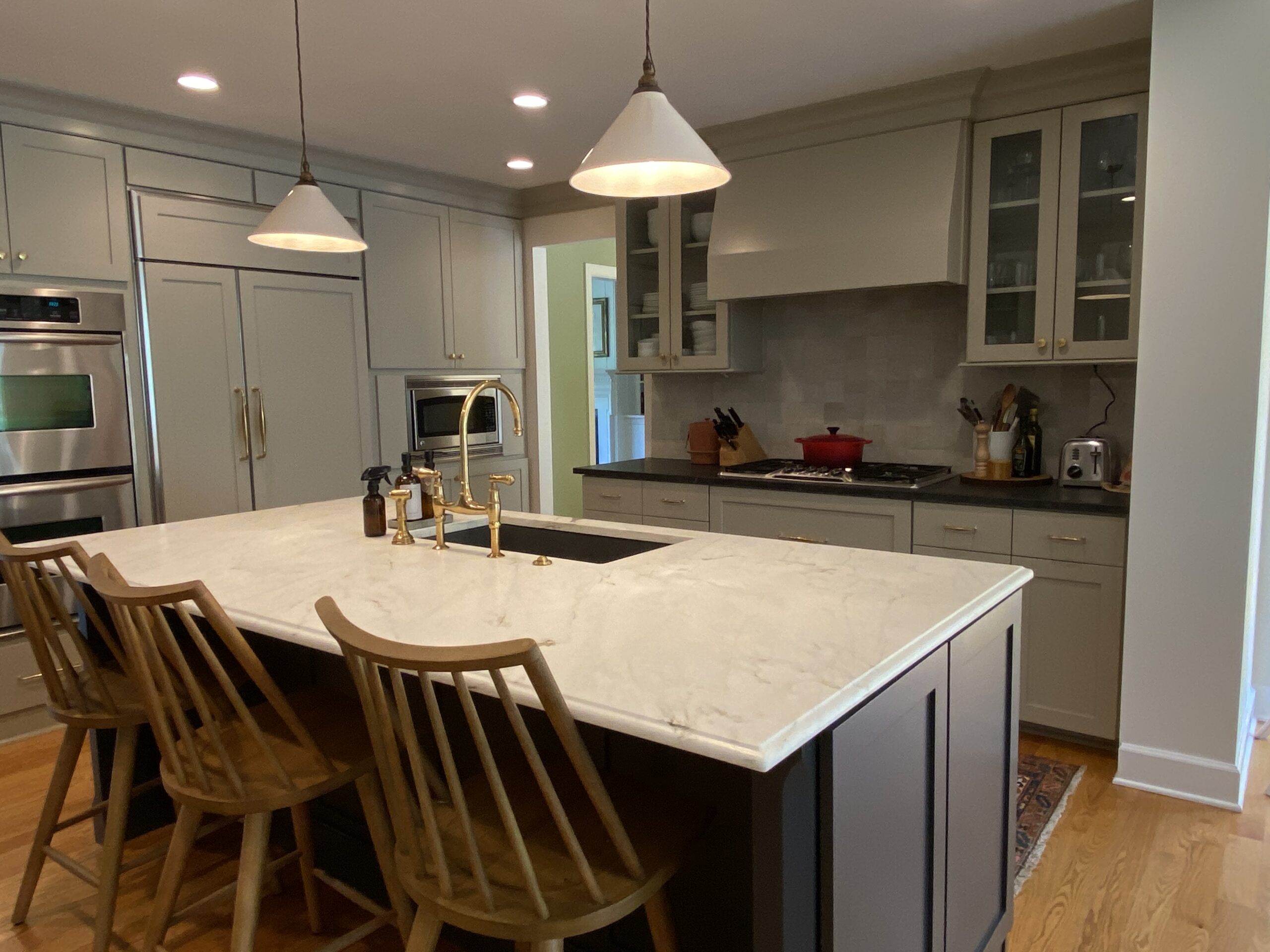 Kitchen with gray cabinetry, marble island countertop, gold fixtures, wood bar stools, stainless steel appliances, and glass-front upper cabinets.