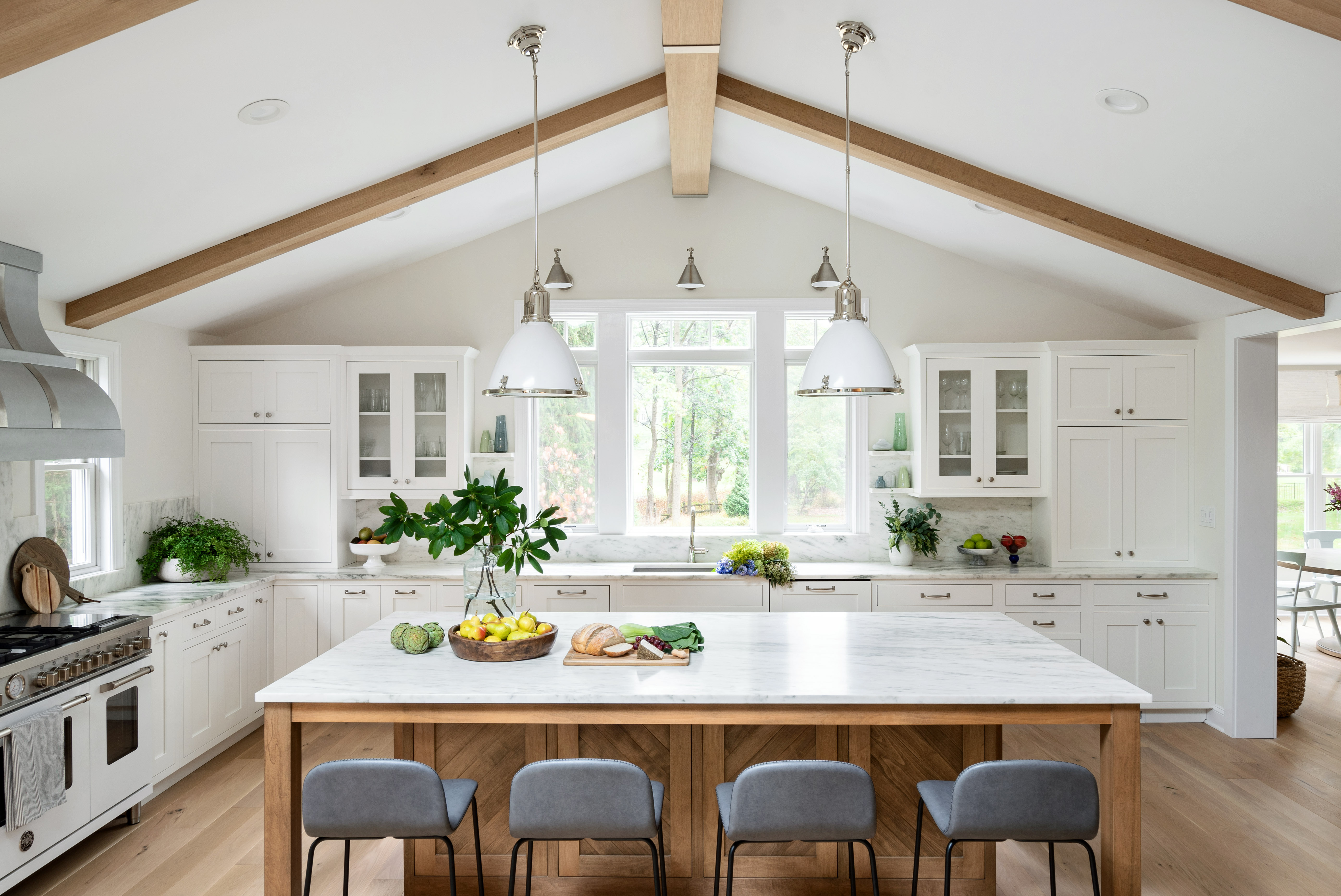 Bright kitchen with vaulted ceiling, wood beams, large island with marble countertop, gray barstools, white cabinetry, and greenery for decor.
