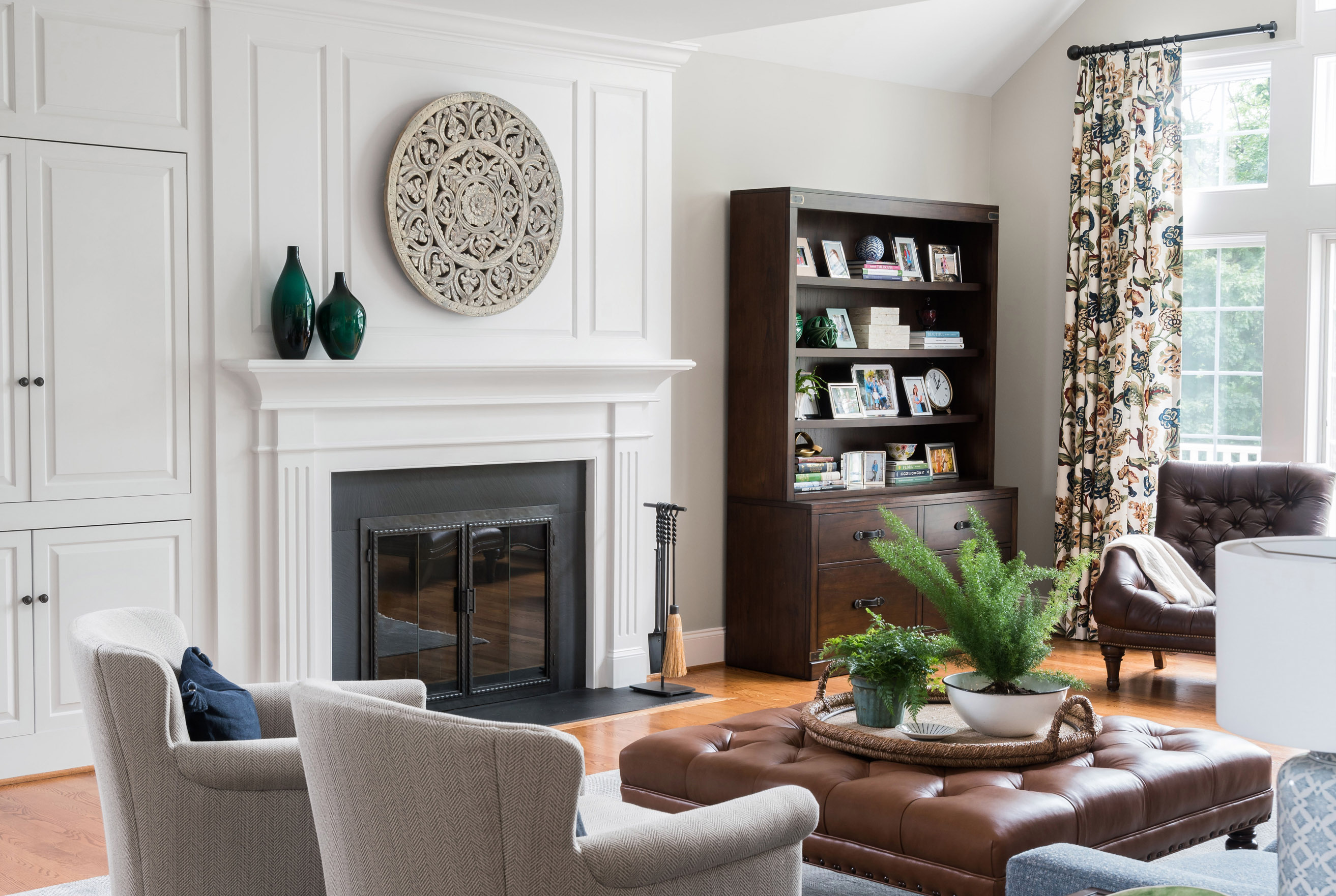 Cozy living room with white fireplace, decorative round wall art, built-in cabinets, dark wood bookshelf, patterned curtains, and leather ottoman with plants.