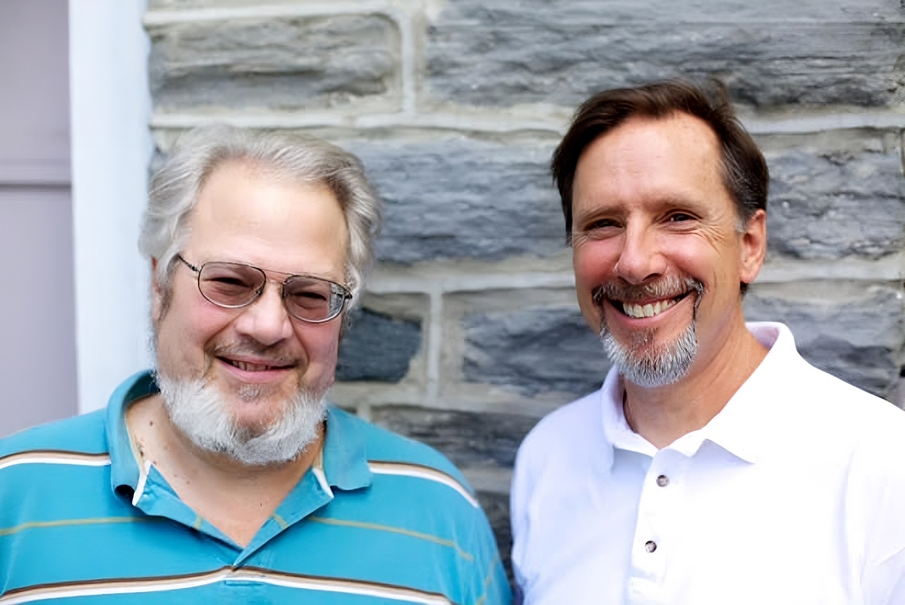 Two men smiling in front of a stone wall. The man on the right is Ted Komacek, owner of TJ's Painting & Renovations.