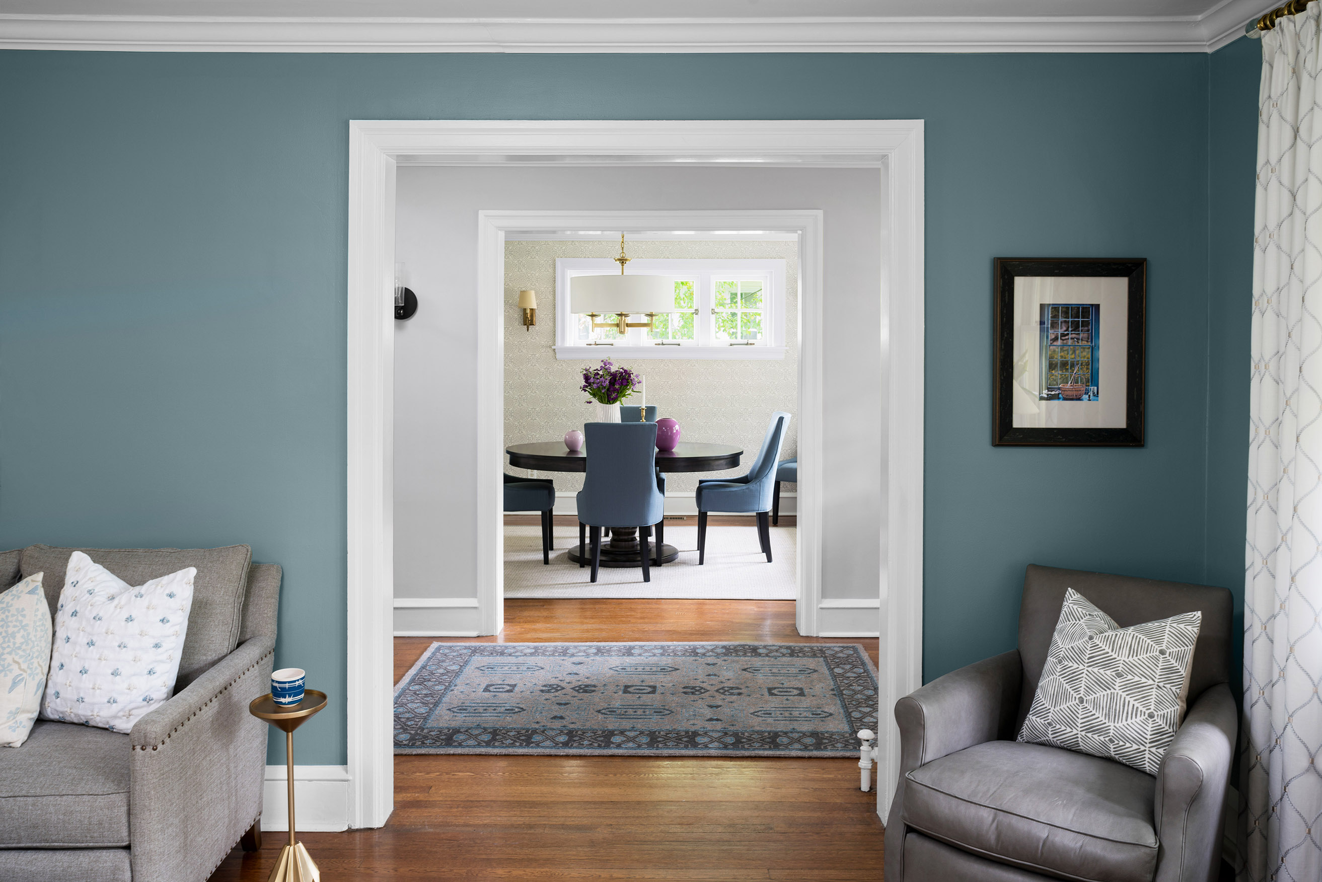 View from a teal living room into a dining room with blue chairs, round table, floral centerpiece, brass light fixture, and patterned area rugs on wood floors.