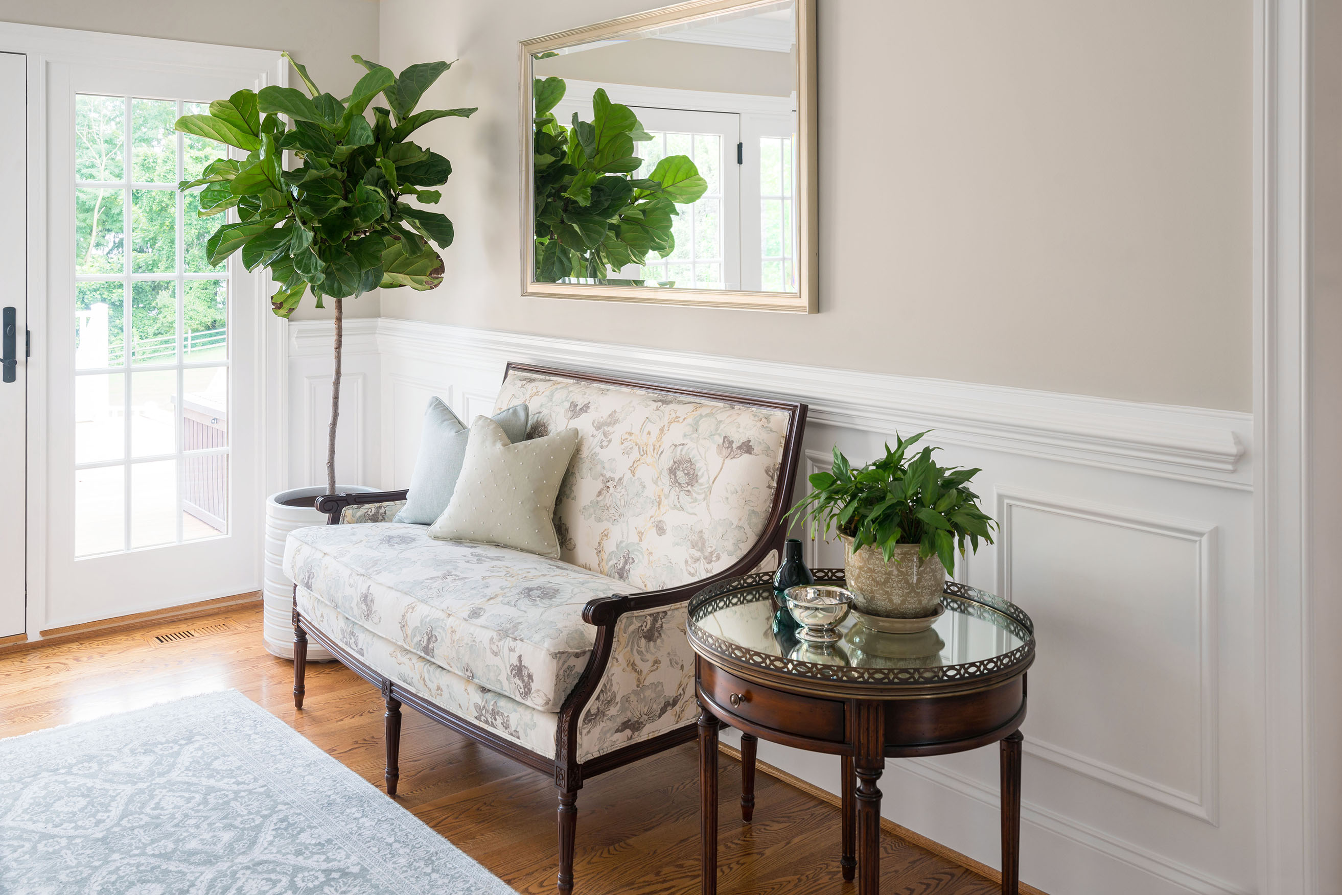 Elegant sitting area with floral upholstered bench, decorative pillows, round glass-top table with potted plant, large mirror, and fiddle-leaf fig by glass doors.