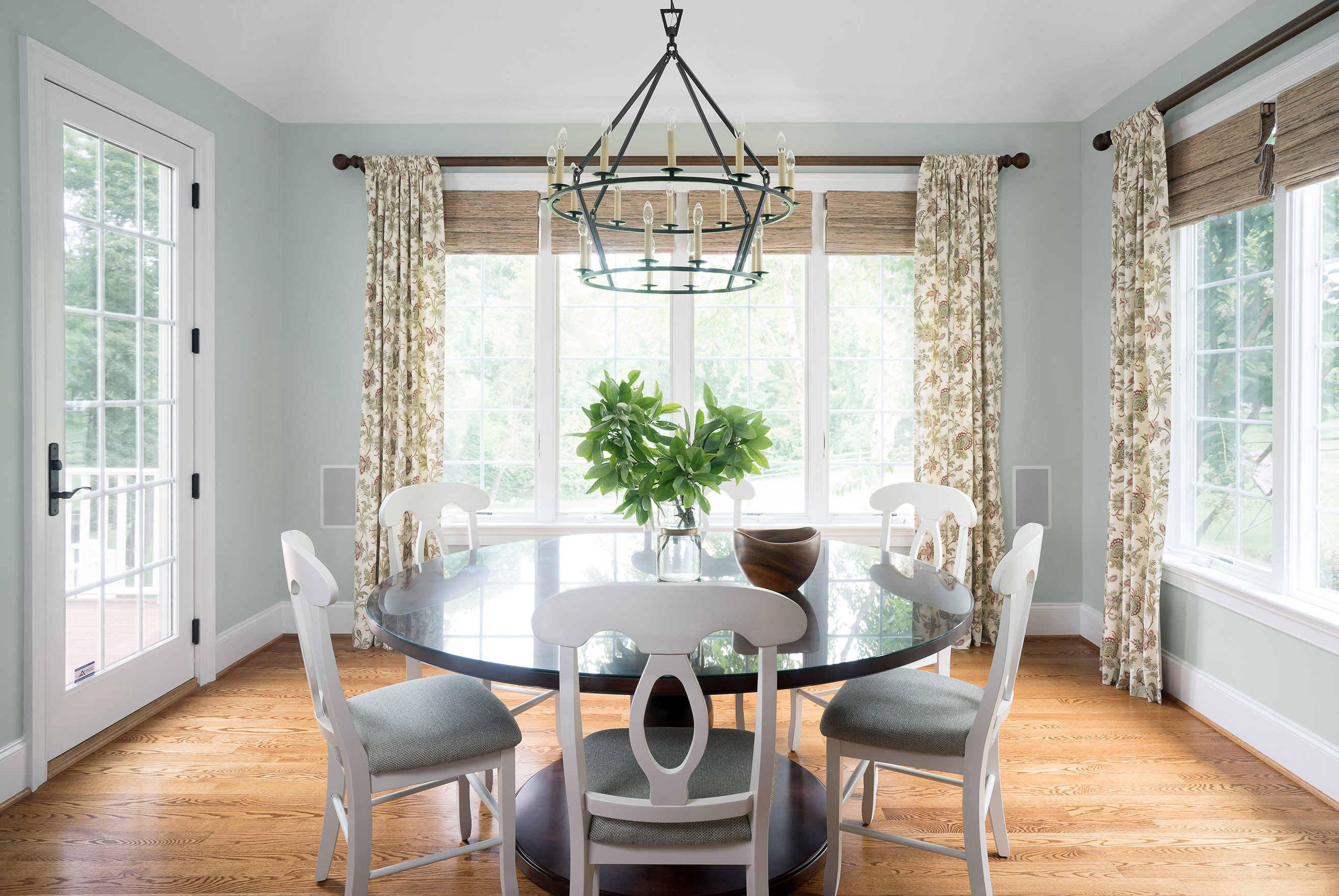Bright dining area with round glass-top table, white chairs, floral curtains, bay windows, black iron chandelier, and vase of greenery as centerpiece.