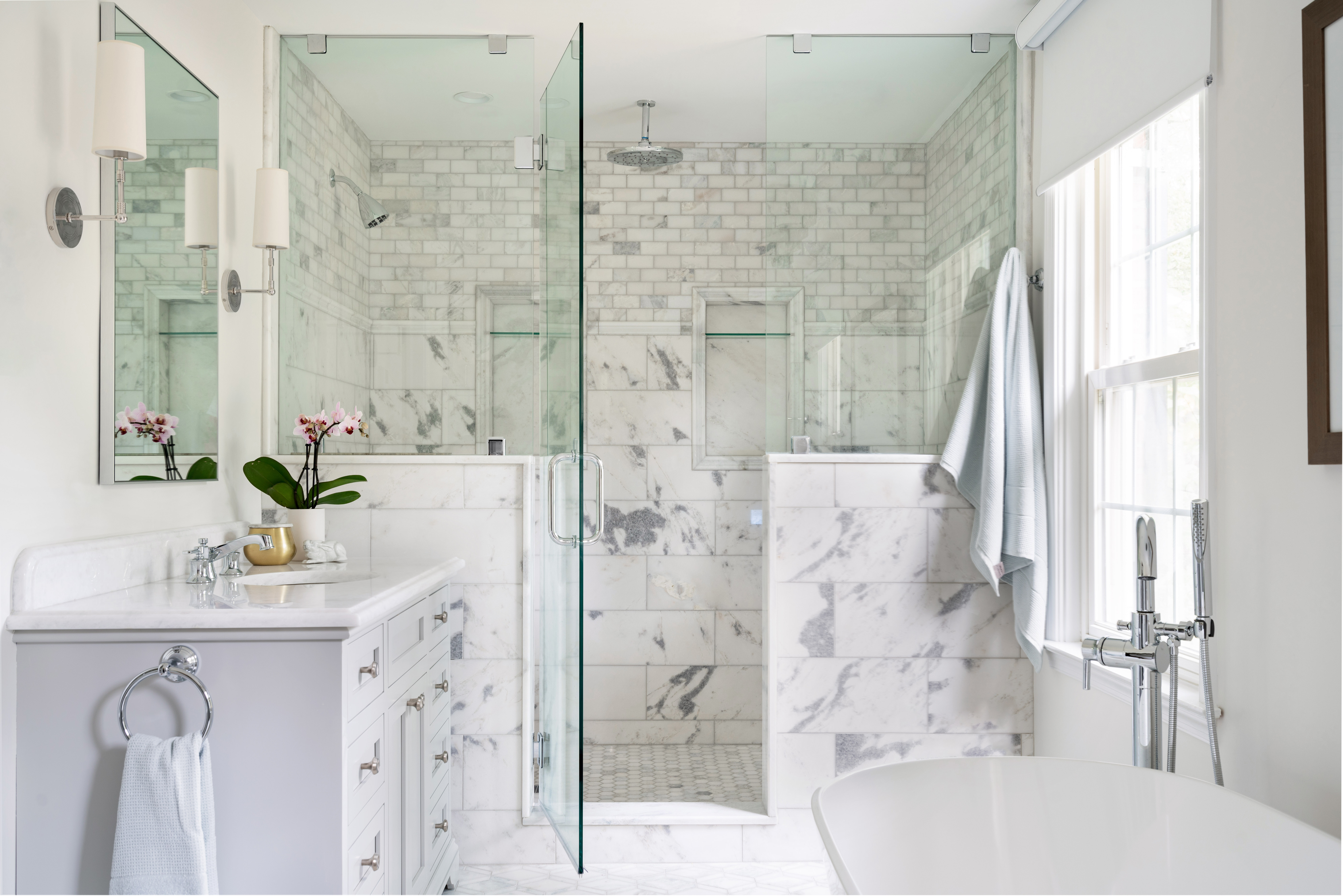 Bright bathroom with glass-enclosed marble shower, double shower heads, white vanity with orchid plant, freestanding tub, and large window with towel hanging.