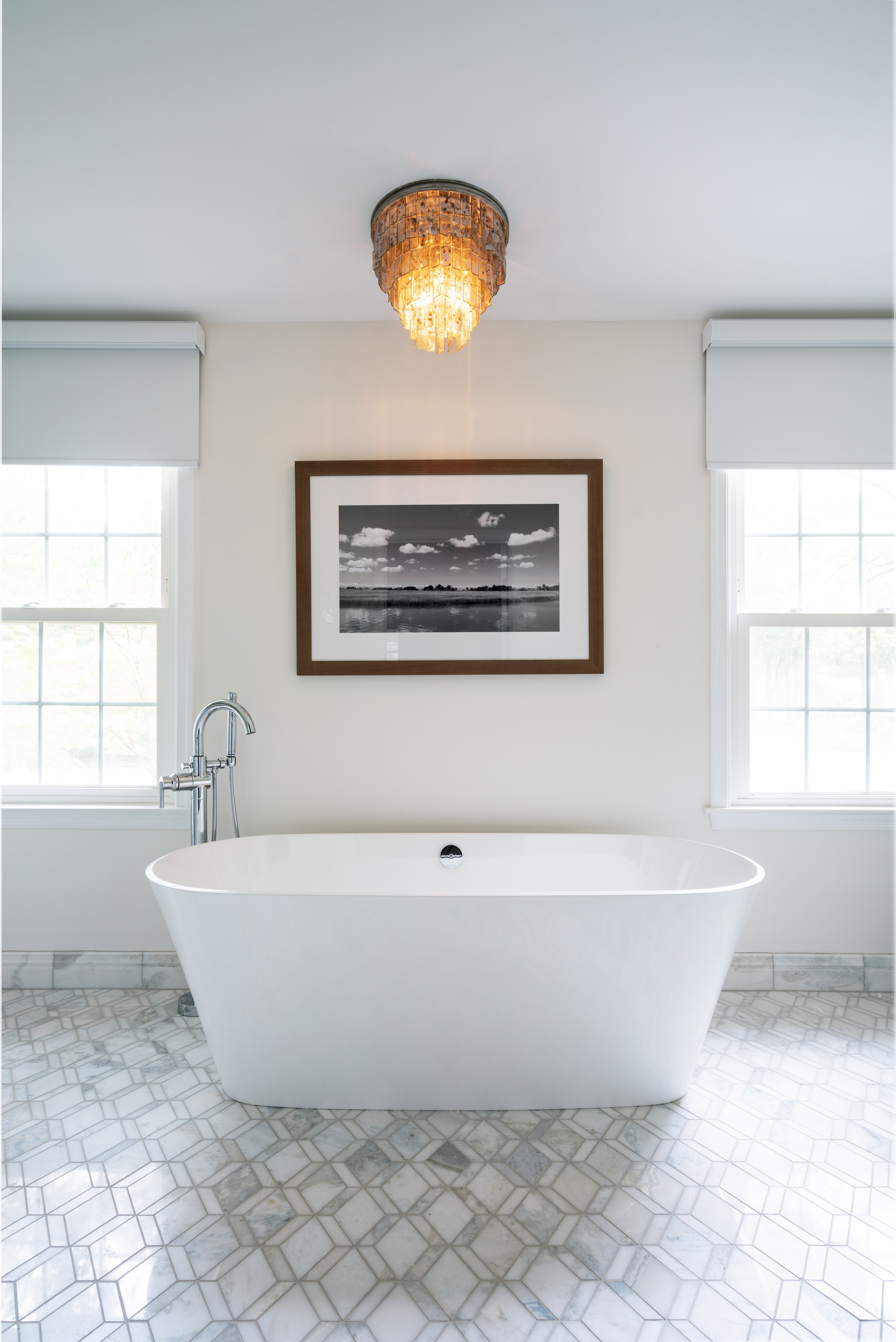 Modern bathroom featuring a freestanding white tub, geometric marble tile floor, chandelier light fixture, and black-and-white framed photo on the wall.