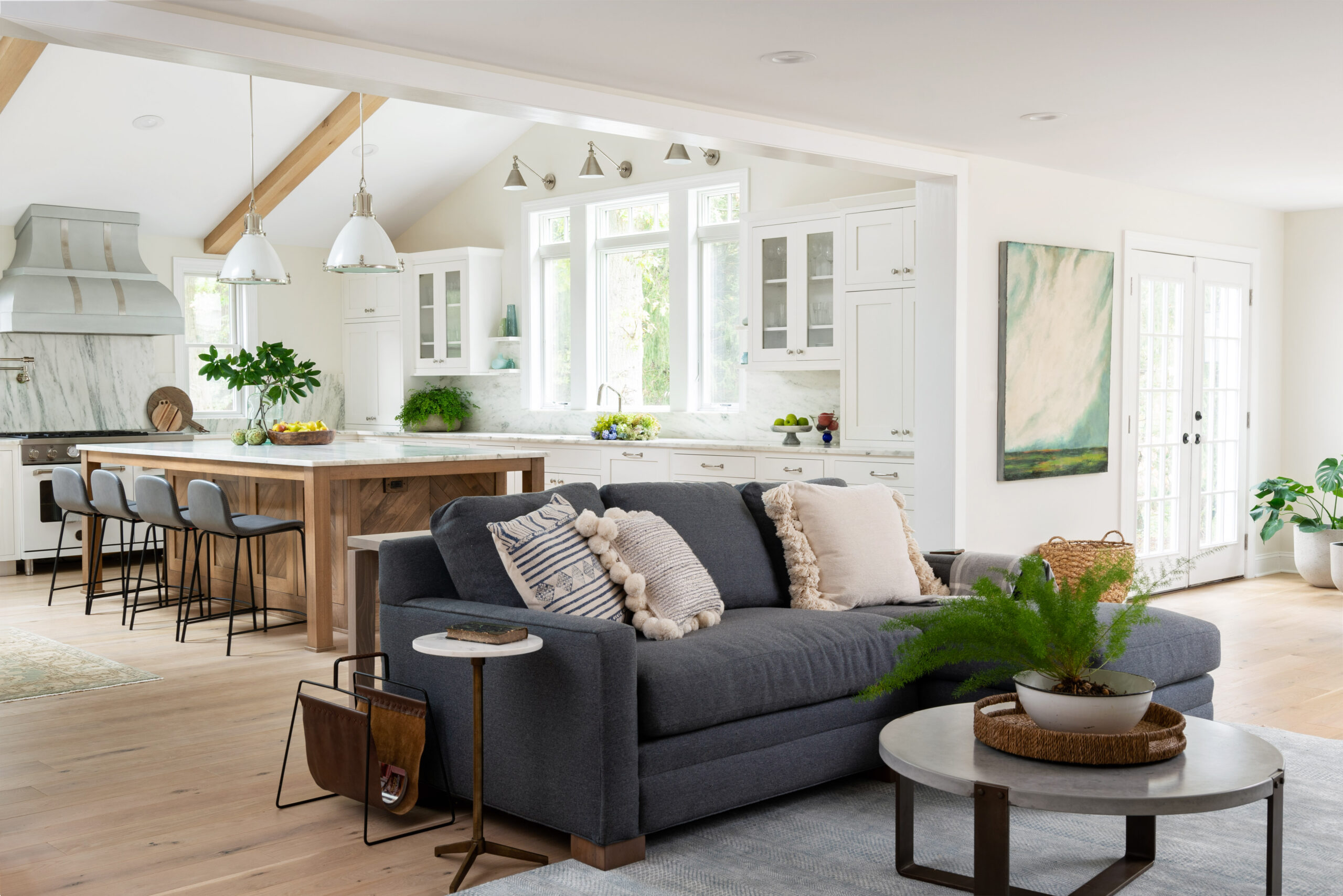 Bright open-concept kitchen and living room with white cabinetry, wood island, gray sofa with pillows, round coffee table, and large windows for natural light.