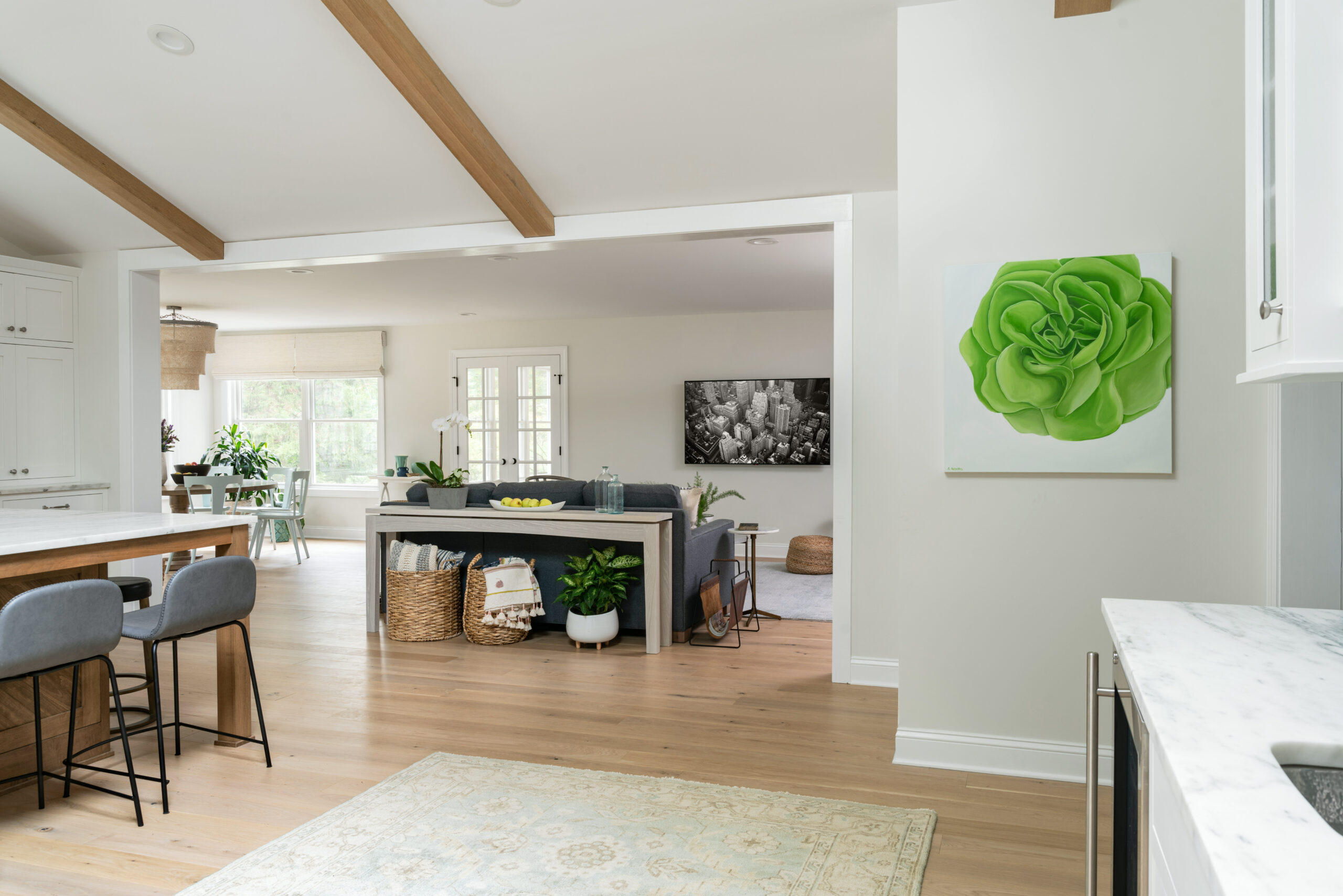 Open-concept kitchen and living space with wood beams, island seating, gray sofa, baskets for storage, large windows, and green rose artwork on the wall.