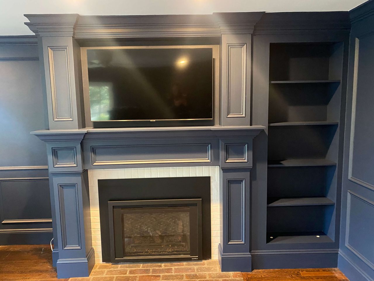 Dark blue built-in fireplace wall with crown molding, a mounted TV above the mantel, white tile fireplace surround, brick hearth, and open shelving.