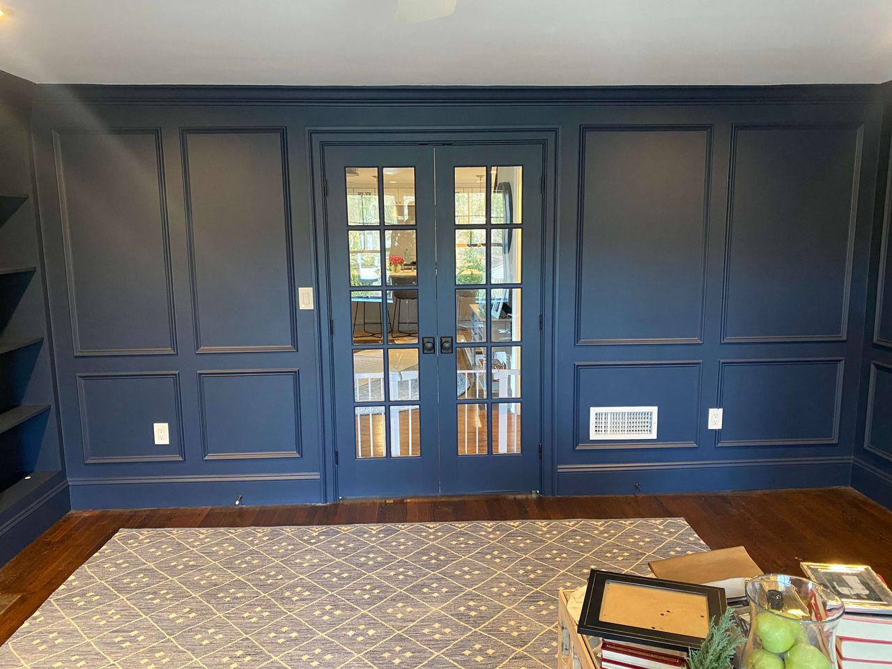 Room with dark blue paneled walls, glass double doors, built-in shelves on the left, hardwood floors, and a patterned rug in front of a small stack of decor.