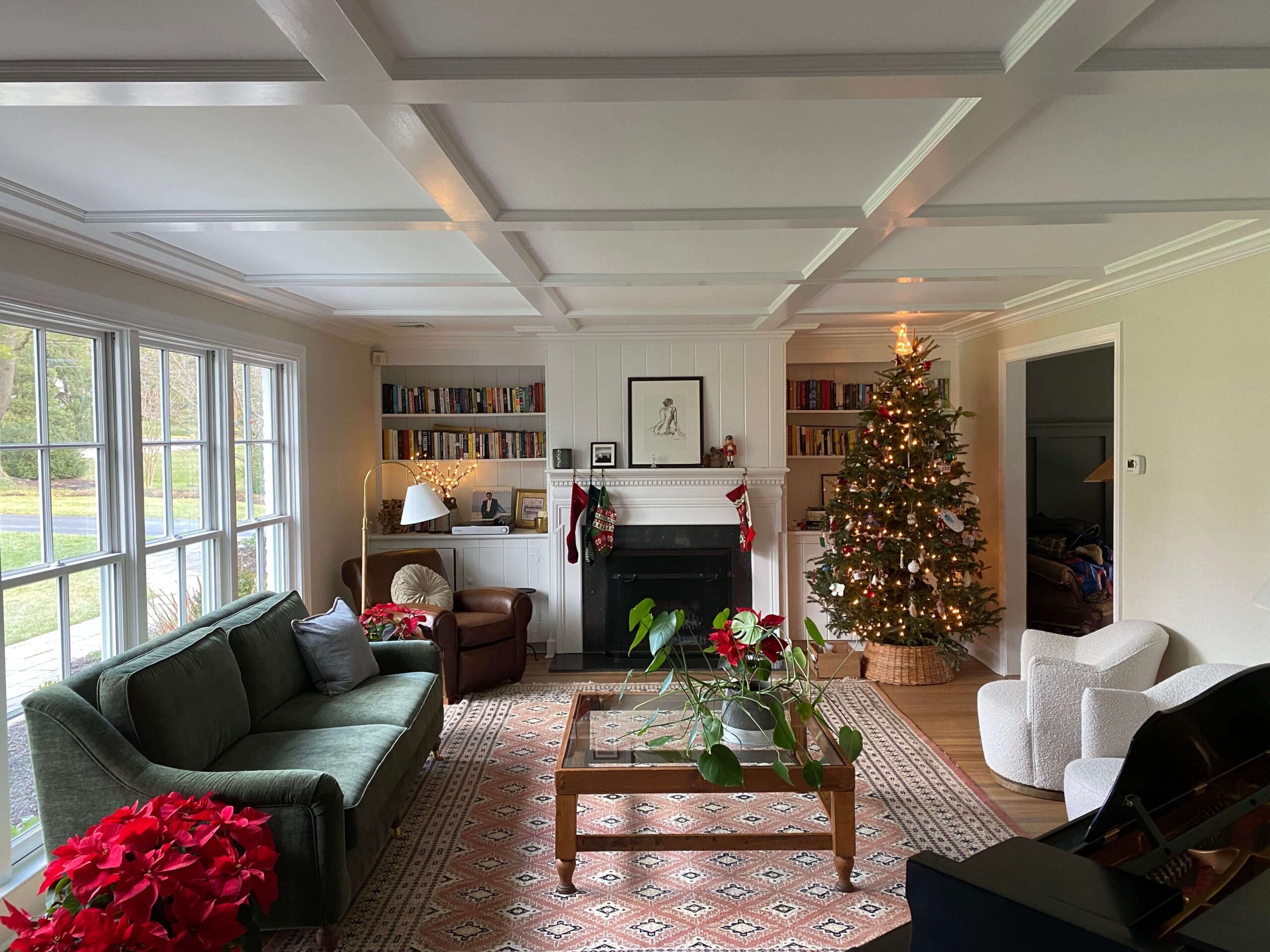 Living room decorated for Christmas with a lit tree, green sofa, armchairs, bookshelves, large windows, and a patterned rug centered under a coffee table.