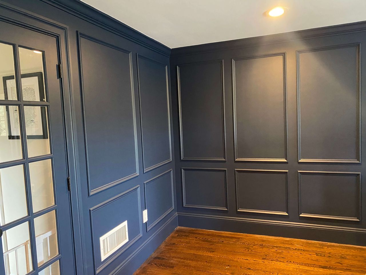 Room with dark blue paneled walls, wood flooring, recessed lighting, and a French door with glass panes leading to another space with framed artwork.